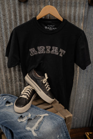 Black t-shirt with 'KRISS' print, sneakers, and jeans on a wooden crate against a corrugated metal background.