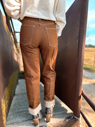 Person wearing brown jeans and a light-colored sweater standing on a rustic metal surface with a blurred natural background.