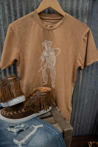 Brown t-shirt with cowboy graphic and fringed shoes on a wooden crate against a corrugated metal background