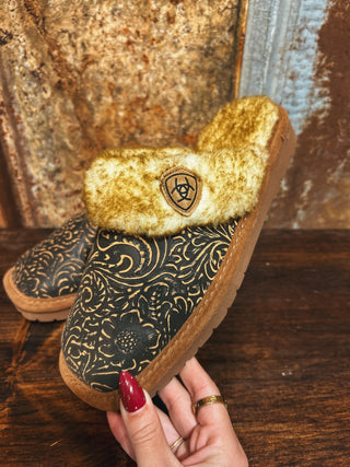 Pair of patterned slippers with a gold interior held by a hand against a rustic wooden background.