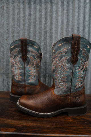 Pair of brown and blue cowboy boots with intricate designs on a wooden surface and metal background.