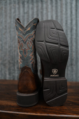 Brown cowboy boot with patterned top and Ariat logo on sole against a rustic background