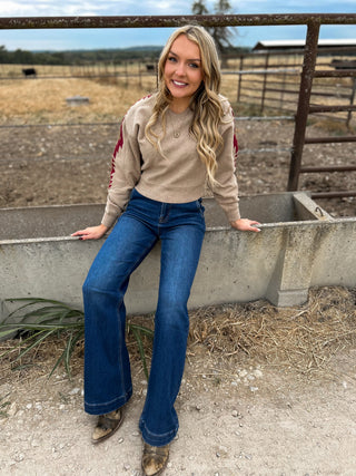 Woman in a beige sweater and blue jeans sitting on a concrete structure in a rural setting.