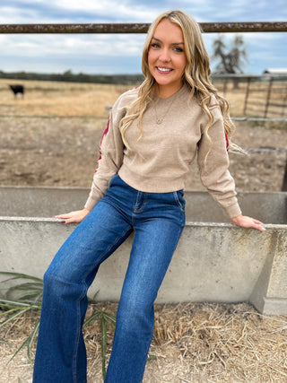 Woman in a beige sweater and blue jeans sitting on a concrete structure with a rural background.
