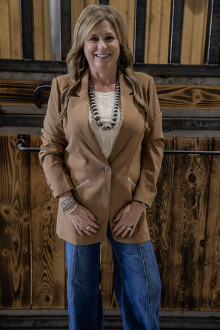 Woman wearing a brown blazer and blue jeans standing in front of wooden panels.
