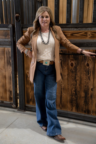 Woman in a beige jacket and blue jeans standing against a rustic wooden wall.