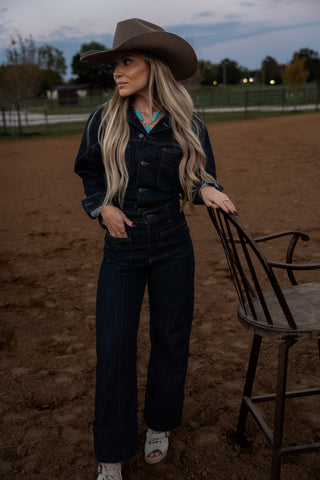 Woman in a cowboy hat and dark outfit standing in a dirt area with a chair.