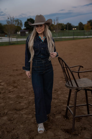 Woman in cowboy hat and dark outfit walking with a chair in an outdoor setting