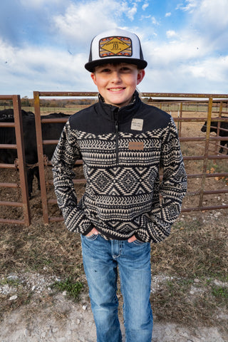 Child wearing a patterned sweater and cap in a rural setting with cows and a fence.