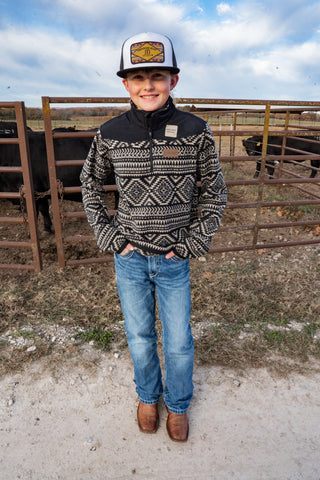 Child wearing a patterned sweater and jeans standing in a fenced area with cows.