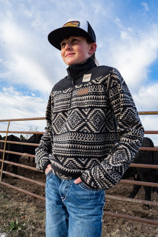 Person wearing a patterned sweater and cap standing in front of a fence with cows.
