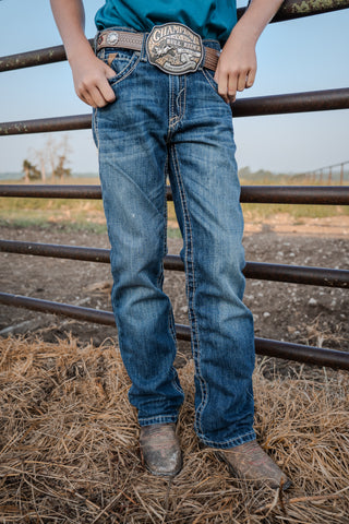 Person wearing blue jeans and a belt with a visible brand logo, standing near a metal fence.