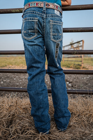 Person wearing blue jeans with a patterned belt standing in front of a metal fence.