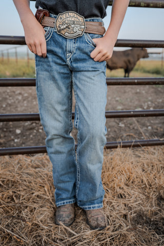 Person wearing blue jeans with a belt and cowboy boots standing near a metal fence with a cow in the background.