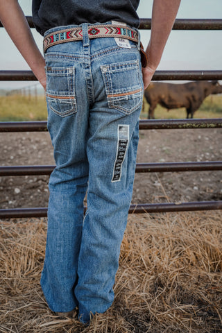 Person wearing blue jeans with a decorative belt, standing in a field with a cow in the background.