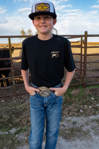 Young boy wearing an Ariat shirt and cap, standing in a rural setting with a fence and open field.
