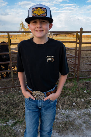 Young boy wearing a black t-shirt with an Ariat logo, blue jeans, and a cap on a farm.