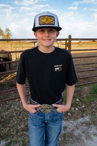 Person wearing a black Ariat t-shirt and blue jeans with a cap, standing in a rural setting.