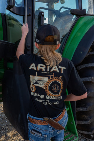 Person wearing an Ariat t-shirt standing next to a large green tractor.