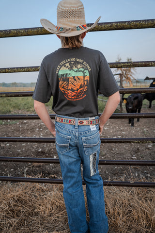 Person wearing a cowboy hat and graphic t-shirt standing in a rural setting with a cow in the background.