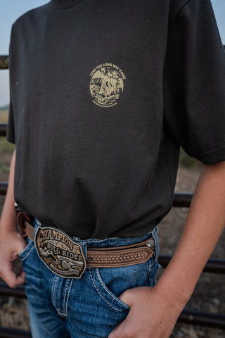 Person wearing a black t-shirt with a logo and blue jeans with a belt, standing outdoors.