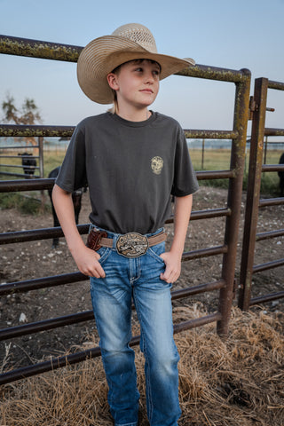 Young boy in a cowboy hat and western attire standing near a metal fence.