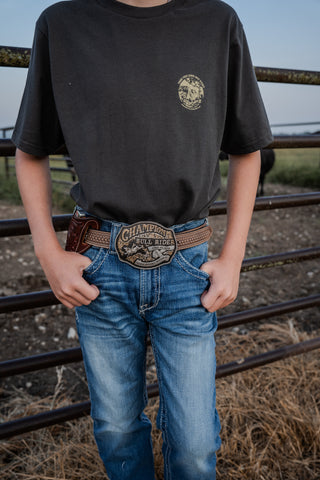 Person wearing a black t-shirt with a logo and blue jeans with a belt, standing in a fenced area.