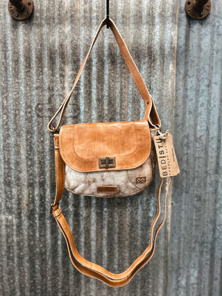 Brown leather handbag with a visible brand tag hanging on a corrugated metal wall.