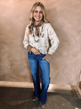 Woman wearing a patterned blouse, blue jeans, and brown boots against a beige wall.