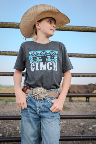 Child wearing a Cinch shirt and cowboy hat with a fence and cow in the background
