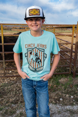 Child wearing a light blue t-shirt with 'Cinch Jeans' logo and a cap, standing in a fenced area.