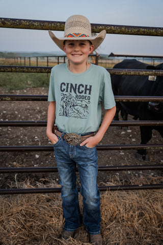 Child wearing a Cinch Rodeo shirt and cowboy hat standing in front of a metal fence with cows in the background.