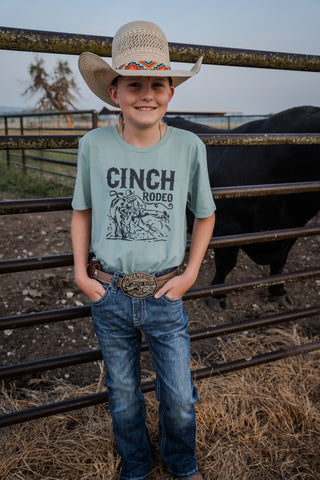 Child wearing a Cinch Rodeo shirt and cowboy hat standing next to a cow in a fenced area.