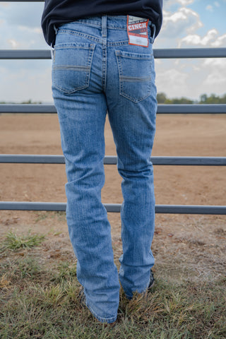 Person wearing blue jeans with a brand tag, standing in front of a metal fence with a rural landscape in the background.