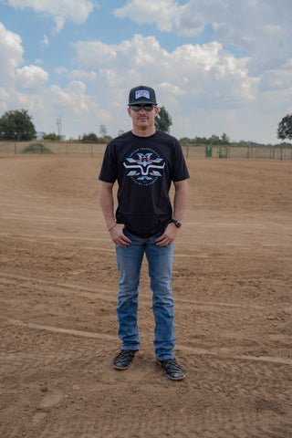 Man wearing a black t-shirt with a logo and blue jeans standing on a dirt field with trees and sky in the background.