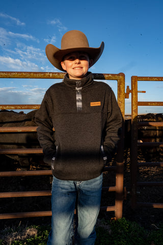 Person wearing a cowboy hat and dark jacket standing in front of a wooden gate with a blue sky background.