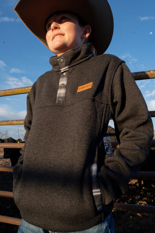 Person wearing a brown jacket with a brand logo, standing outdoors against a blue sky.