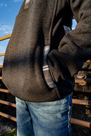 Person wearing a brown jacket and blue jeans standing behind a wooden fence with a blue sky.