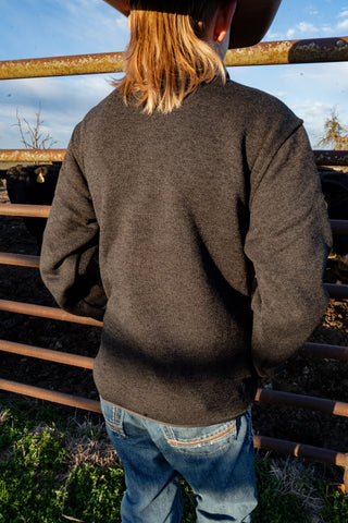 Person wearing a brown jacket and blue jeans standing behind a metal fence with a blue sky and trees in the background.