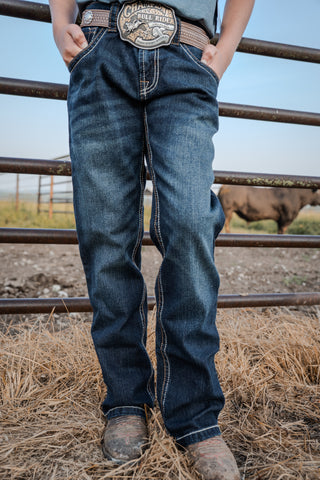 Person wearing blue jeans with a belt buckle standing in front of a metal fence with a cow in the background.
