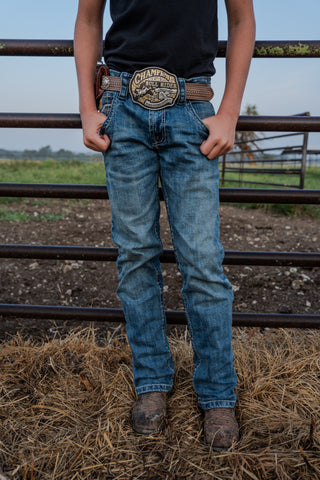 Person wearing blue jeans and a belt with a prominent brand logo, standing in a field.
