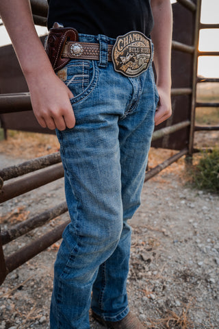Person wearing blue jeans with a belt and patch, standing in front of a metal gate.