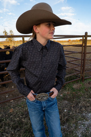 Person wearing a cowboy hat and patterned shirt standing in a rural setting with a fence and open field.