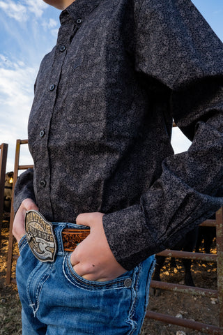 Person wearing a dark button-up shirt and blue jeans with a belt buckle, standing outdoors.