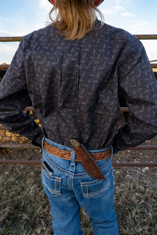Person wearing a dark jacket and blue jeans with a belt and leather pouch, standing in an outdoor setting.