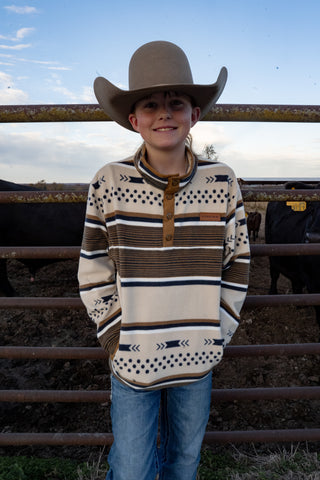 Person wearing a cowboy hat and patterned shirt standing in front of a fence with cows in the background.
