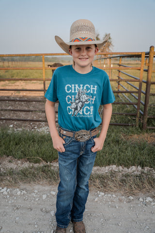 Young boy in a cowboy hat and blue jeans standing in front of a wooden fence with a horse in the background.