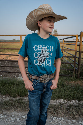 Child wearing a blue t-shirt with 'Cinch' branding, standing in front of a wooden fence.