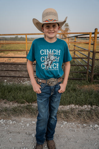 Young boy in a cowboy hat and blue jeans standing on a dirt path with a wooden fence and grassy area in the background.