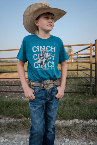 Young boy in a teal shirt with a horse graphic and 'Chinch' text, wearing a cowboy hat and belt, standing in a rural setting.